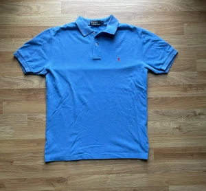 Blå pikétröja från Polo Ralph Lauren - Snygg blå pikétröja från Polo Ralph Lauren i storlek M. Tröjan har klassisk krage, två knappar och den ikoniska röda broderade loggan på bröstet. Tillverkad i mjuk bomull som känns skön mot huden. Perfekt för dig som gillar stilren och enkel design.