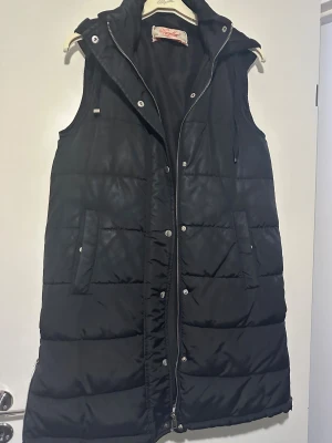 Svart lång väst med huva - Svart väst i fint skick! Några ljusare partier från användning men inget som stör. Lite öppen söm strax innan luvan – superenkelt att fixa!
