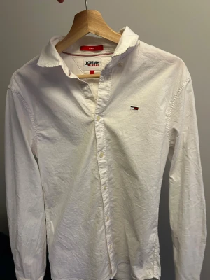 Vit skjorta från Tommy Jeans slim fit - Vit skjorta från Tommy Jeans i slim fit-modell med klassisk krage och knappar framtill. Liten broderad logga på bröstet och diskret logodetalj på ryggen. Skjortan är långärmad och tillverkad i mjuk bomull som känns skön mot huden.