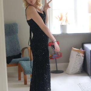 Svart glittrig långklänning med slits - svart långklänning med glittriga paljetter över hela klänningen. Klänningen har smala axelband, figurnära passform och en hög slits på sidan som gör den extra fin. Perfekt för bal eller fest när du vill sticka ut och glittra.