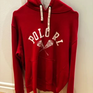 Röd hoodie från Polo Ralph Lauren - Snygg röd hoodie från Polo Ralph Lauren med vit text och lacrosse-klubbor på bröstet. Tröjan har huva med vita snören och en stor magficka. Perfekt för chill dagar och streetstyle. Långärmad och i mjukt material.