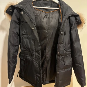 Svart dunjacka med fuskpäls från Hollies - Svart dunjacka från Hollies med stor huva och snygg fuskpäls i bruna toner. Jackan har flera fickor med lock, dragkedja och tryckknappar framtill. Perfekt för kalla dagar med sin varma och fluffiga känsla. Köpt för ungefär 4000 kr, och knappast använd, därför vill jag sälja den. Pris kan diskuteras!