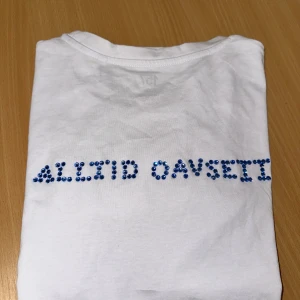 Vit t-shirt med blå paljetter på ryggen  - Storlek XS Vit t-shirt med texten 'ALLTID OAVSETT' baktill. Klassisk rund hals och korta ärmar. Tröjan i grund är från lager 157, går att tvätta OBS! Bara i 30 grader, ingen torktumling 