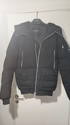 Svart pufferjacka Supply & Demand - Svart pufferjacka från Supply & Demand med avtagbar fuskpäls till huvan. Jackan har flera fickor med dragkedja, ribbade muddar och en snygg patch på ärmen. Perfekt för kalla dagar och har en modern, urban vibe. Litet hål innefrån jackan men inget skadligt som påverlar alls 