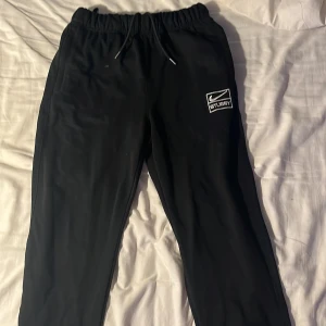 Svarta Stussy x Nike mjukisbyxor S - Svarta mjukisbyxor från Stussy x Nike med broderad logga på vänster ben. Byxorna har elastisk midja med snörning och är gjorda i mjukt bomullsmaterial. Perfekt för chill eller streetwear och har en loose passform.