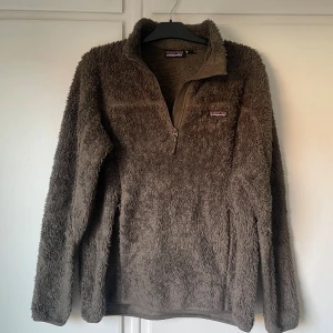 Olivgrön fleece-tröja från Patagonia | M - Mjuk och fluffig brun fleece-tröja från Patagonia i storlek M. Tröjan har lång ärm, halv dragkedja och hög krage. Perfekt för kyliga dagar och har en chill, avslappnad vibe. Patagonia-logga på bröstet. Snyggt och bekvämt plagg för höst och vinter.