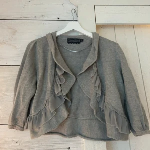 Grå kofta med volang  - Supersöt grå kofta från Vintage Stories i storlek M. Koftan har volangdetaljer framtill och längs nederkanten, samt en mjuk, stickad känsla. Perfekt för dig som gillar romantiska och chill vibes. Långärmad och med en snygg krage som ger extra stilpoäng.