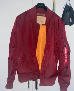 Vinröd bomberjacka Alpha Industries - Säljer en vinröd bomberjacka från Alpha Industries i storlek S. Jackan har klassisk 'Remove Before Flight'-tag på ärmen, ribbade muddar och dragkedja framtill. Tillverkad i slitstark polyester med orange foder och flera fickor. Perfekt för dig som gillar streetwear.
