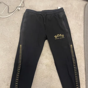 Svarta mjukisbyxor från Hugo Boss - Svarta mjukisbyxor från Hugo Boss med guldfärgade detaljer längs benen och logga på vänster lår.