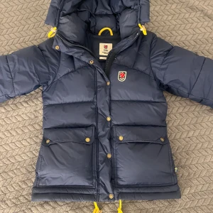 Blå dunjacka från Fjällräven - Fortfarande osäker beroende på pris så skriv om du har intresse! KÖP INTE KONTAKTA FÖRST!! blå dunjacka från Fjällräven med huva, stora fickor och snygga gula detaljer. Jackan har dragkedja och knappar framtill samt klassisk Fjällräven-logga på bröstet. Perfekt för kalla dagar och riktigt skön att bära. Jackan är i xxs sitter jätte bra på mig defekter ser ni på bilderna men de är inget som märks av, Skriv för mer bilder!