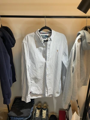 Randig skjorta Polo Ralph Lauren M - Snygg slim fit skjorta från Polo Ralph Lauren i vitt och blått med tunna ränder. Klassisk krage, knappar framtill och broderad logga på bröstet. Långärmad och tillverkad i mjuk bomull. Perfekt för dig som gillar stilrena och tidlösa plagg.