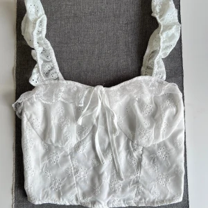 Vit broderad topp - Superfin vit topp med broderade blommor och spetsdetaljer. Axelbanden är breda och har volanger. Framtill en söt snörning och tyget är lätt och luftigt. Baksidan är smockad för extra komfort. Aldrig använd.