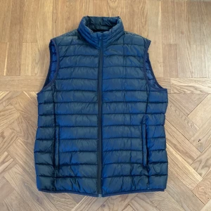 Mörkblå dunväst med dragkedja - Snygg svart dunväst med marinblå insida och hög krage. Västen har hel dragkedja framtill och två fickor på sidorna. Perfekt att slänga över en hoodie eller tröja för extra värme. Enkel och stilren look som funkar till det mesta.