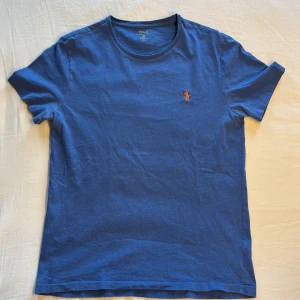 Blå t-shirt från Polo Ralph Lauren - Snygg blå t-shirt från Polo Ralph Lauren i custom fit. Klassisk rund hals. På bröstet finns den ikoniska orangea Polo-loggan broderad. Tillverkad i mjuk bomull som känns skön mot huden. Perfekt för en clean och stilren look. Priset går att diskuteras vid snabbaffär!