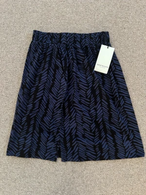 Blåmönstrade shorts från Hofmann - Snygga shorts från Hofmann Copenhagen med blått och svart abstrakt mönster. Modellen är loose fit med elastisk midja för skön passform. Materialet känns lätt och luftigt, perfekt för varma dagar. Stilsäkert val för dig som gillar grafiska prints.                                                     Viktiga funktioner  Mörkblå casual kjol från Hofmann Copenhagen  A-linjeformad design  Unikt fiskbensmönster  Knälång längd  100% bomull  Storlek 36  Ny med taggar 💕