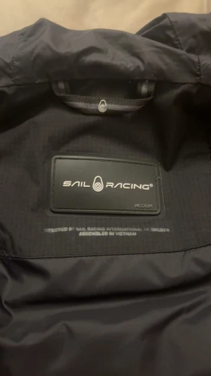 Svart Sail Racing dunjacka med huva - Svart dunjacka från Sail Racing, modell Antarctica Expedition. Jackan har huva, dragkedja framtill, logga på bröstet och ärmen samt flera praktiska fickor. Perfekt för kalla dagar och har en clean, sportig look.