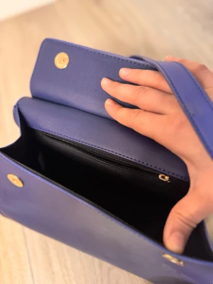 Blå axelväska med gulddetaljer - Snygg blå axelväska i skinnimitation med stilrena gulddetaljer. Väskan har både handtag och justerbar axelrem, samt magnetlås och innerfack med dragkedja. Modern och boxig form som ger en cool vibe till din outfit.