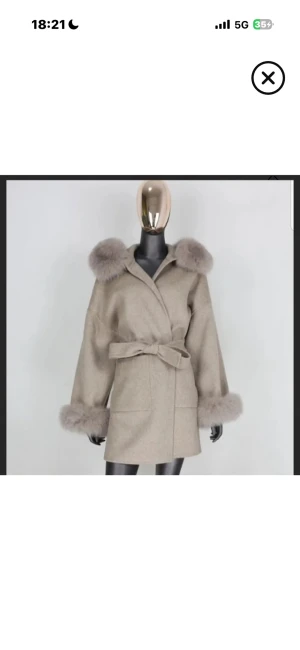 Beige kappa med pälsdetaljer och bälte - Säljer en beige kappa med fluffiga pälsdetaljer vid krage och ärmslut. Jackan har en omlottmodell med bälte i midjan och breda, långa ärmar. Perfekt för dig som vill ha en mysig och stilren look under höst och vinter.