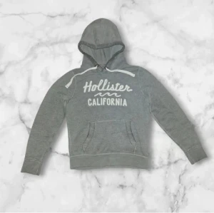 Grå hoodie från Hollister  - Snygg grå hoodie från Hollister California med vit textprint på bröstet. Tröjan har huva med snörning, känguruficka och ribbade muddar vid ärmslut och nederkant. Perfekt för chill dagar och streetstyle.