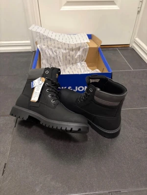 Svarta boots från Jack & Jones - Säljer ett par svarta boots från Jack & Jones med grov sula och snörning. Skorna har rund tå, grå detaljer vid skaftet och är tillverkade i syntetmaterial med nubuck-look. Perfekt för dig som gillar robust stil och vill ha något som funkar hela hösten och vintern.