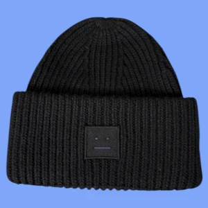 Svart ribbad mössa Acne Studios - Svart ribbstickad mössa från Acne Studios med klassisk uppvikt kant och det ikoniska Face-logotypen framtill. Mjuk och varm, perfekt för kyliga dagar. Snyggt minimalistisk design som passar till allt.