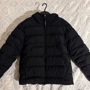 Svart pufferjacka från C.P. Company - Svart pufferjacka från C.P. Company med ikoniska goggles i huvan. Jackan har dragkedja framtill, två stora fickor och en skön, vadderad känsla. Perfekt för kalla dagar och har en cool streetwear-vibe. Huvan är justerbar och jackan är riktigt varm. 