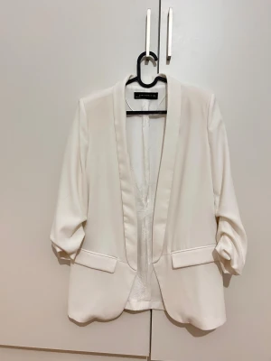 Vit kavaj från Zara Woman, M - Snygg vit kavaj från Zara Woman i storlek M. Stilren design med klassisk krage, två fickor framtill och lätt uppvikta ärmar. Materialet är mjukt och har en lätt glansig finish som ger en fräsch look. Perfekt för att lyfta din outfit.