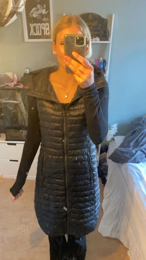 Lång svart dunväst från Everest - Säljer en lång, svart dunväst från Everest med huva och dragkedja hela vägen. Västen är quiltad och har en snygg, figurnära passform. Perfekt att slänga över en hoodie eller tröja för extra värme. Riktigt stilren och enkel att matcha.