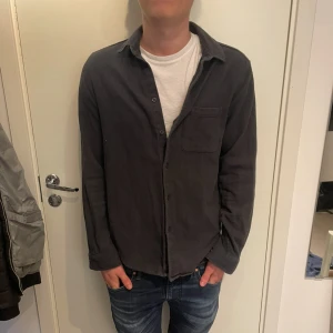 Overshirt  - Snygg mörkgrå skjorta från Samsøe Samsøe i mjukt tyg med klassisk krage och bröstficka. Skjortan har knäppning framtill och långa ärmar, perfekt för en avslappnad men stilren look.