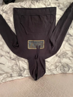 Svart hoodie från Emporio Armani EA7 - Svart hoodie från Emporio Armani EA7 med stort logotryck i grått och guld på bröstet. Tröjan har huva med snörning och en klassisk magficka. Mjuk insida och ribbade muddar vid ärmslut och nederkant. Perfekt för dig som gillar stilrena märkesplagg.