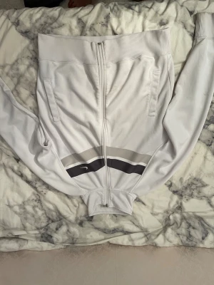 Vit vindjacka från Nike med ränder - Säljer en fräsch vit vindjacka från Nike med coola svarta och gråa ränder över bröstet. Jackan har hel dragkedja, ribbad krage och muddar samt två sidofickor. Perfekt för dig som gillar sportig stil och vill ha något som sticker ut.