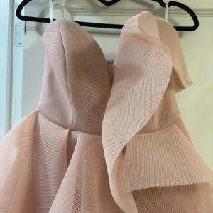  volangklänning - Säljer en rosa (lite åt gammelrosa/pudderrosa) klänning med flera lager volanger och en asymmetrisk design. Klänningen är axelbandslös och har en snygg omlott-detalj framtill. Materialet är lätt och ger ett exklusivt intryck, perfekt för dig som vill sticka ut med en unik look.