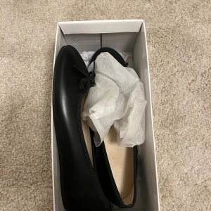 Svarta ballerinaskor med rosett - Stilrena svarta ballerinaskor med rund tå och liten rosett framtill. Skorna har en platt sula och är tillverkade i ett slätt material som ger en klassisk look. Dom är oanvända alltså i nytt skick