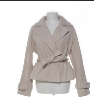 Nelly Jacka Beige omlottjacka med bälte - Snygg beige jacka med omlottknäppning och brett bälte i midjan. Jackan har bred krage, långa ärmar med dekorativa spännen vid ärmsluten och en lite kortare, figurnära passform. Perfekt för dig som gillar stilrena och trendiga plagg.