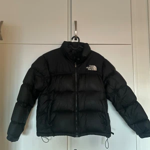 Svart pufferjacka från The North Face - Säljer en svart pufferjacka från The North Face i storlek M. Jackan har hög krage, broderad logga på bröstet och ryggen, samt dragkedja framtill. Fylld med dun och har snörning i nederkant för extra passform. Perfekt för kalla dagar.