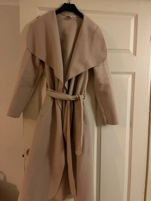 Beige omlottkappa från NLY Trend - Snygg beige kappa från NLY Trend i omlottmodell med bred krage och bälte i midjan. Jackan har långa ärmar och praktiska fickor på sidorna. Perfekt för dig som gillar stilrena och trendiga ytterplagg med en chill vibe.