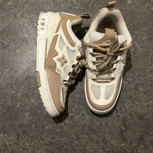 LV skates Sneaker - Säljer ett par Louis Vuitton sneakers i beige och vitt med LV-logga och blommig detalj på sidan. Skorna har rund tå, platt sula och grova snören. Materialmix av skinn, mocka och mesh ger en lyxig vibe. Säljes då de inte används och är i toppskick.