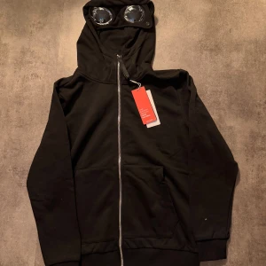 Svart hoodie med goggles från C.P. Company - Svart hoodie från C.P. Company med ikoniska goggles i huvan och broderad logga på bröstet. Tröjan har dragkedja framtill, känguruficka och ribbade muddar. Materialet är mjuk bomull som känns skön mot huden. Perfekt för dig som gillar streetwear och unika detaljer. Råkat tvättas från storlek L nu passar den S😭