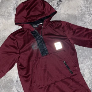 Vinröd Nike Air Max hoodie med half zip - Snygg vinröd hoodie från Nike Air Max med huva och half zip framtill. Stor ficka med dragkedja på magen och svart Air Max-detalj längs dragkedjan. Tillverkad i mjuk polyester, perfekt för chill eller träning. Ikonisk Nike Air Max-logga på bröstet.