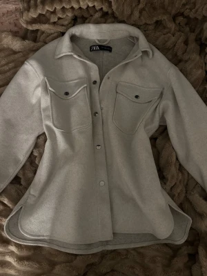 Beige overshirt från Zara - Snygg beige overshirt från Zara med klassisk krage och stora bröstfickor med knappar. Skjortan har långa ärmar och är tillverkad i mjukt ullmaterial. Perfekt för lager-på-lager och har en loose passform med rundad nederkant.
