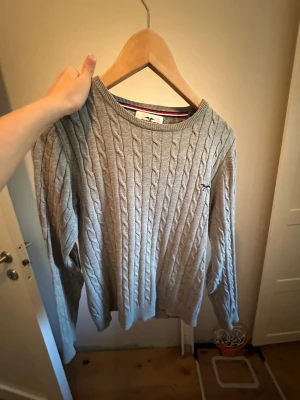 Grå kabelstickad tröja Hampton Republic Gant - Snygg grå kabelstickad tröja från Hampton Republic med rund hals och diskret logga på bröstet. Tröjan har långa ärmar och ribbade muddar vid ärmslut och nederkant. Perfekt för dig som gillar klassisk stil med en modern touch.