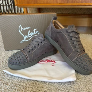 Christian Louboutin grå mocka sneakers - Snygga grå sneakers från Christian Louboutin i mocka med ikoniska nitar på tån och klassisk röd sula. Skorna har snörning och en rund tå, vilket ger en cool och edgy vibe. Perfekta för dig som vill sticka ut med exklusiv design och detaljer.