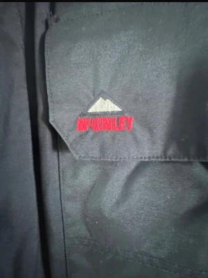 Svart parkajacka från McKinley L - Svart parkajacka från McKinley i storlek L. Jackan har huva med snörning, två stora fickor med lock och broderad logga på bröstet. Perfekt för kalla dagar med sin långa passform och funktionella design. Materialet är slitstark polyester. Mycket fin och varm jacka nu när vintern kommit. Den var dyr i inköp så priset är otroligt bra. 😊 