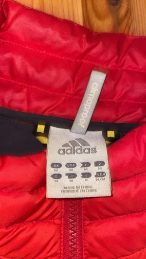 Röd dunjacka från Adidas - Säljer en röd dunjacka från Adidas med klassisk logga på bröstet. Jackan har quiltad design, lång ärm och dragkedja framtill. Materialet är lätt och smidigt, perfekt för kyliga dagar. Insidan är mörkgrå och jackan har en clean, sportig look.