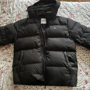 Svart pufferjacka från Tommy Jeans - Svart pufferjacka från Tommy Jeans med huva och dragkedja. Jackan har broderad logga på bröstet och ärmarna, samt stora quiltade sektioner för extra värme. Perfekt för kalla dagar och har en clean, modern look.