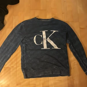 Blå Calvin Klein Jeans sweatshirt - Säljer en blå sweatshirt från Calvin Klein Jeans med stort vitt CK-tryck på bröstet och är en gammal model. Tröjan har rund hals, långa ärmar och ribbade muddar. Den har ett litet hål vid tagen.Perfekt för dig som gillar klassiska streetwear-vibbar och vill ha en snygg logga framtill..