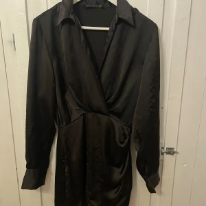 Svart omlottklänning från Zara XS - Snygg svart kort omlottklänning från Zara i satinliknande polyester. Klänningen har lång ärm, klassisk krage och markerad midja. Perfekt för dig som gillar stilrena och eleganta plagg med en modern twist. Passar mig som oftast har S