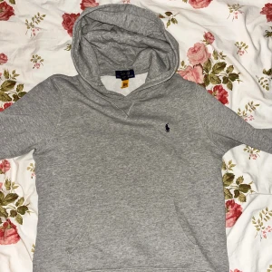 Grå Polo Ralph Lauren hoodie - Säljer en stilren grå hoodie från Polo Ralph Lauren med klassisk huva och känguruficka. Tröjan har lång ärm och är tillverkad i mjuk bomull. Passar nog Xs också. Vid några funderingar tveka inte med att höra av er. Pris går att diskutera vid snabb affär 