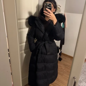 Svart lång dunjacka med päls - Säljer en svart, lång dunjacka från FUR Stockholm med fluffig svart päls på huvan (avtagbar). Jackan har bälte i midjan som är avtagbar och knappar fram till. Storlek S. Helt ny skick. 