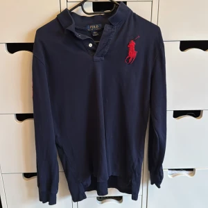 Marinblå pikétröja från Ralph Lauren 14-16 years - Snygg marinblå långärmad pikétröja från Ralph Lauren med stor röd broderad logga på bröstet. Klassisk krage och knappslå framtill. Perfekt för dig som gillar stilrena och sportiga plagg med premiumkänsla. Strl L = 14-16 years